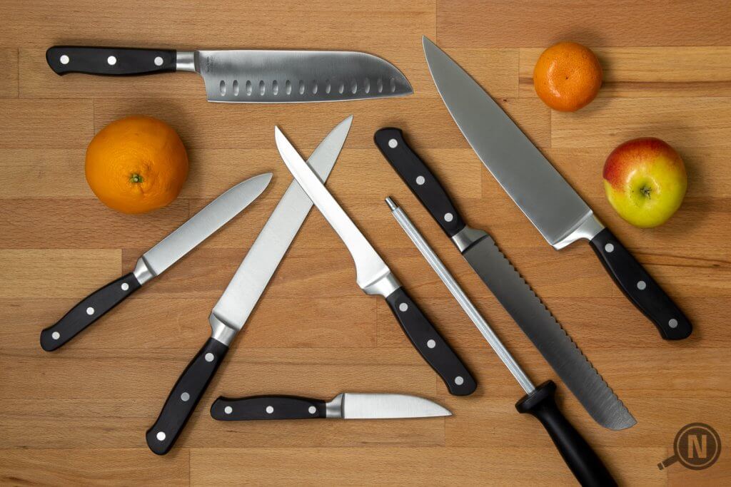 Flatlay mit mehreren unterschiedlichen Messern, dazwichn als Dekoration Orange, Mandarine und Apfel